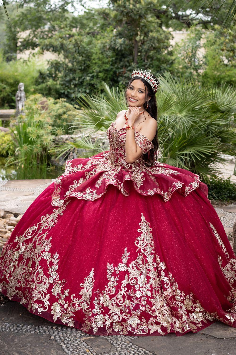 A stunning burgundy and gold quinceañera ball gown with intricate lace appliques photographed in a garden setting.