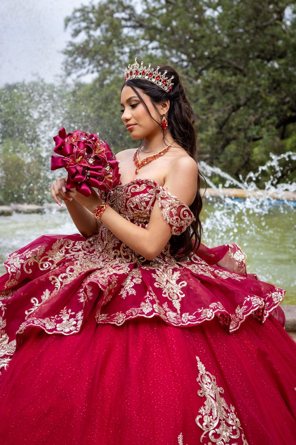 A stunning red and gold quinceañera dress with intricate embroidery details is showcased in an outdoor garden setting.