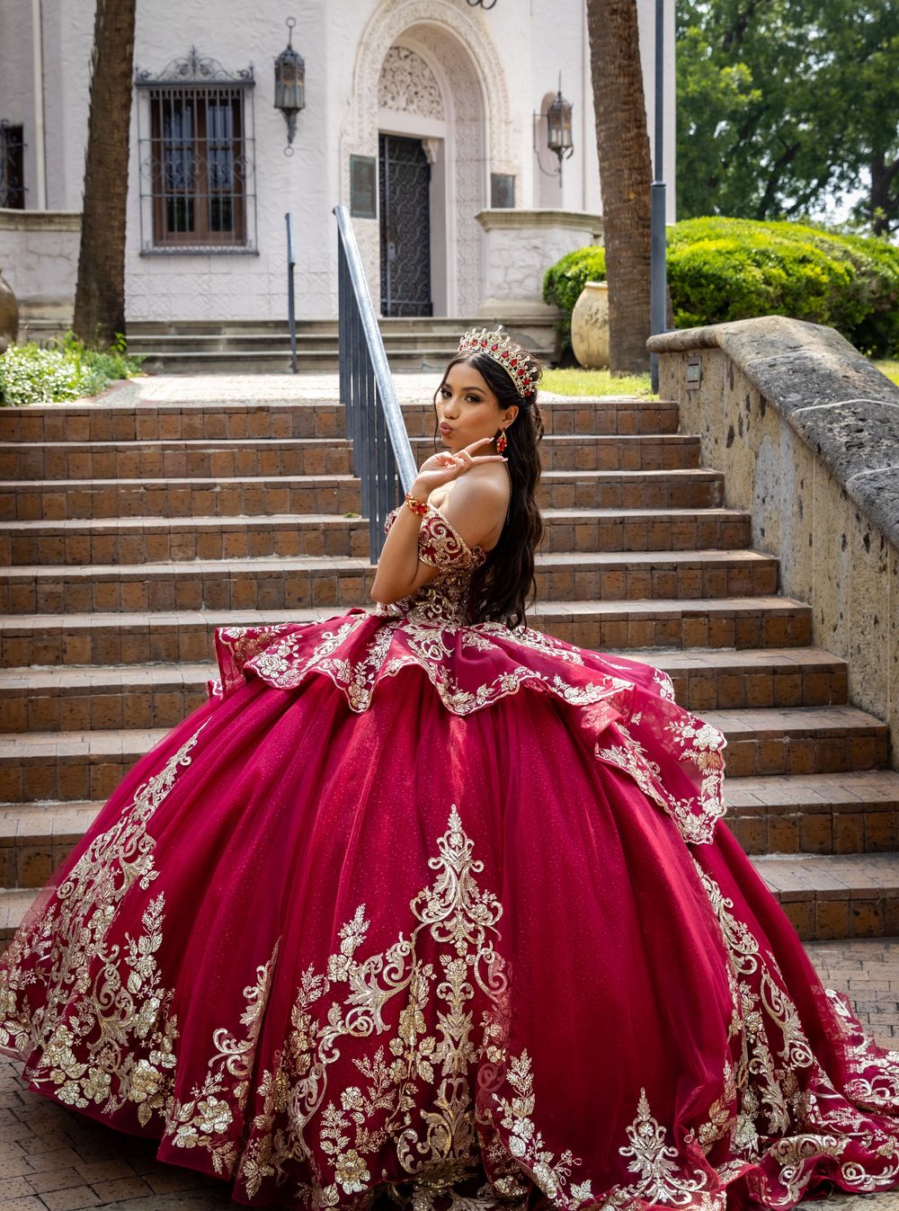 A stunning burgundy and gold quinceañera gown with intricate lace details photographed on stone church steps.