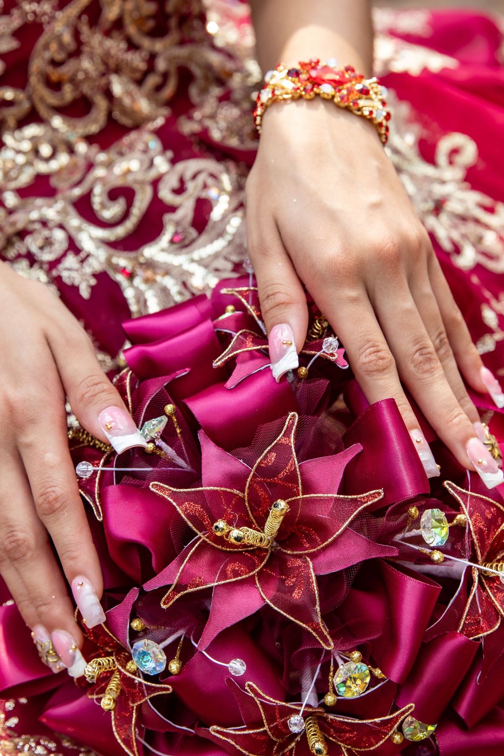 Detailed view of hands with manicured nails holding burgundy fabric flowers adorned with golden wire and crystals.