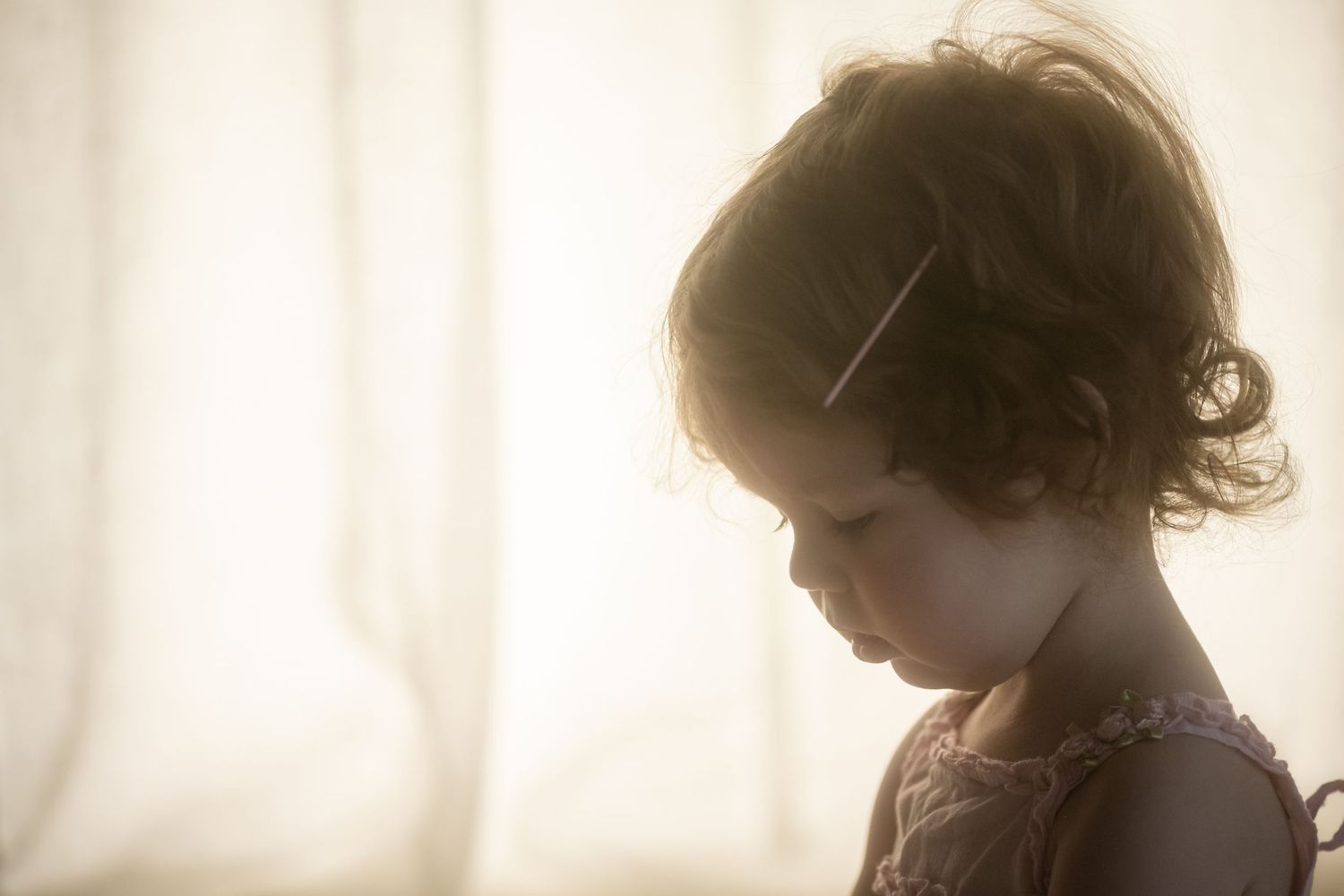 Young girl with curly hair looks down pensively in soft, dreamy backlit portrait photography.