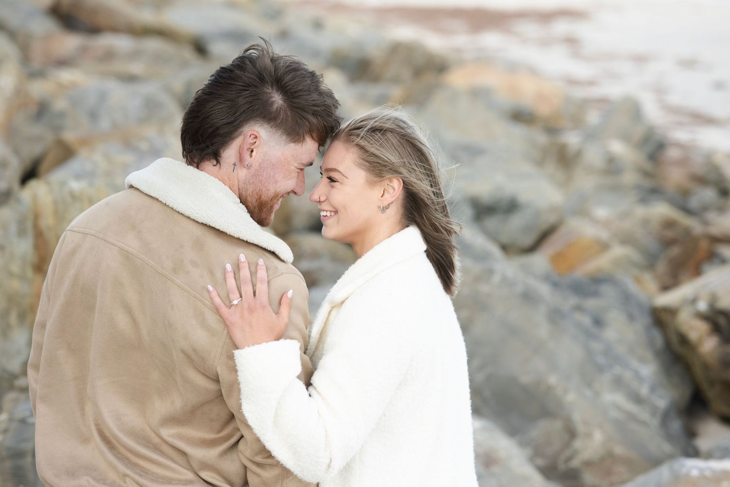 Couple sharing a romantic moment outdoors near rocky shore, woman in white coat smiling at man in tan jacket.