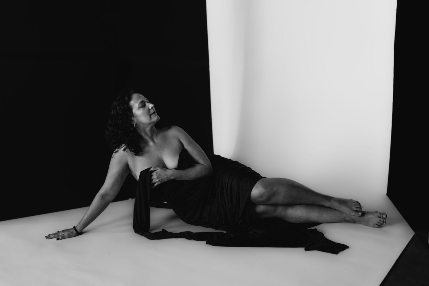 Black and white artistic photo of woman reclining on floor draped in black fabric against white backdrop.