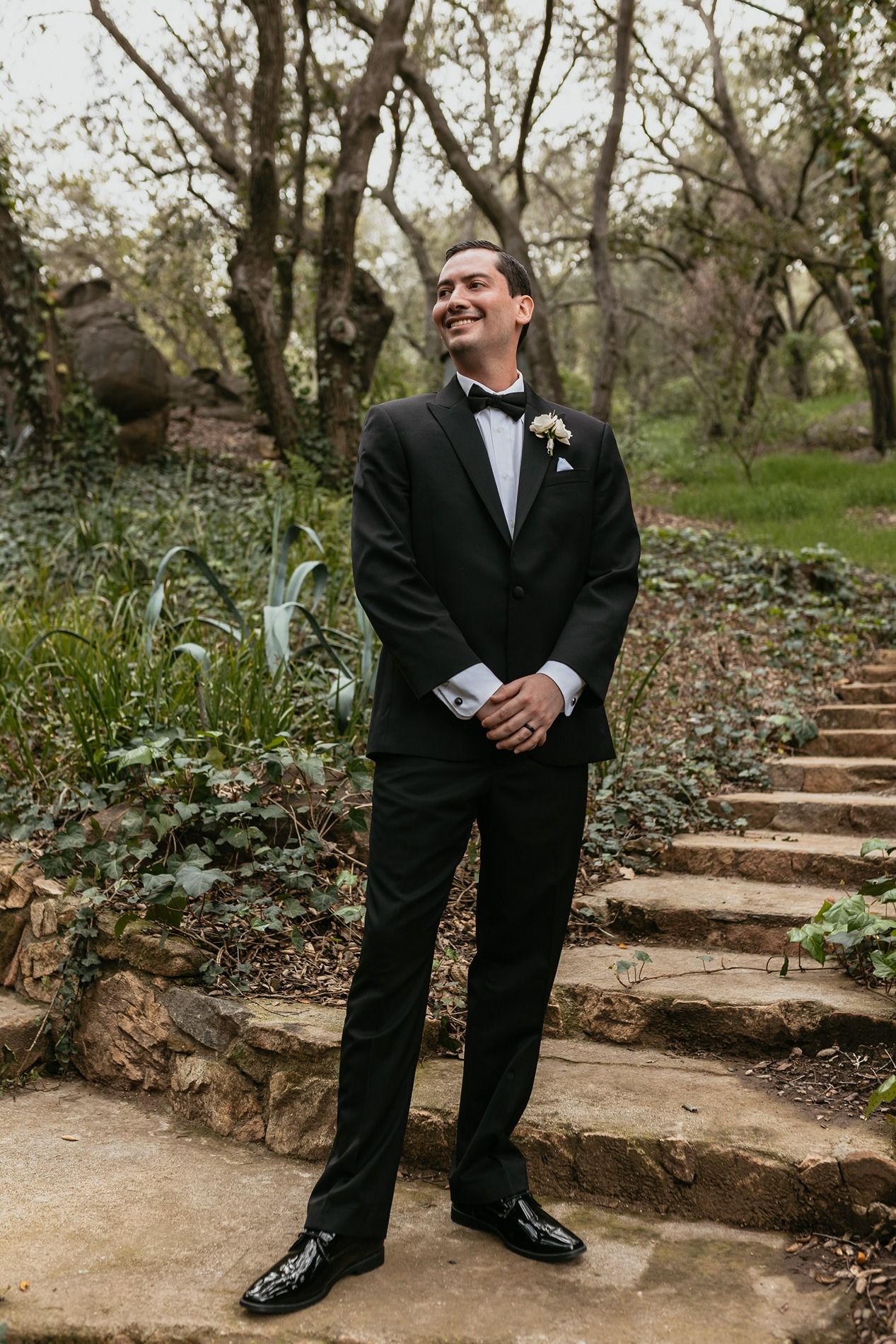 A groom in a black tuxedo standing on stone garden steps in a natural outdoor setting at The Oaks at Duncan Lane.