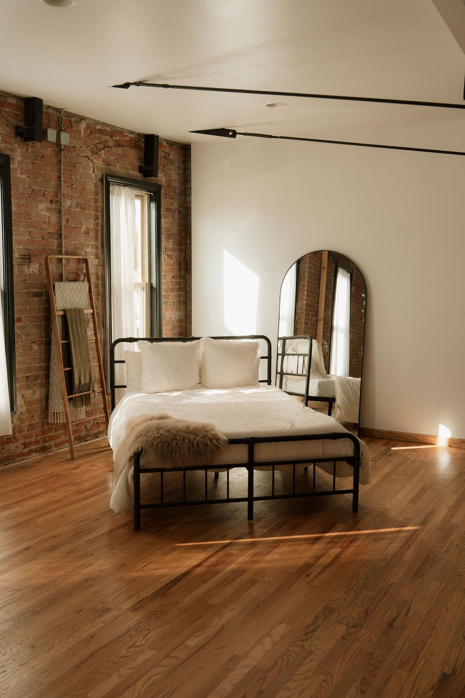Minimalist bedroom with exposed brick walls, hardwood floors, and a simple metal bed frame in warm natural lighting.
