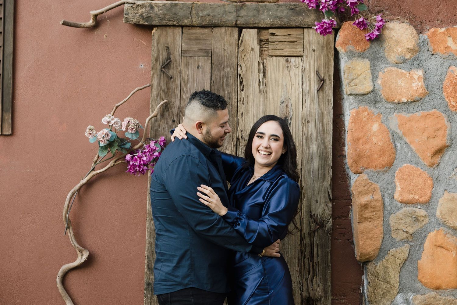 A couple in matching navy jackets share a romantic moment against a rustic wooden door and stone wall.