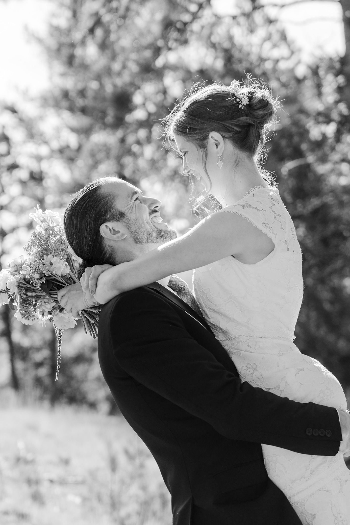Bride holding colorful bouquet stands with smiling groom in suit among trees at outdoor fall wedding.