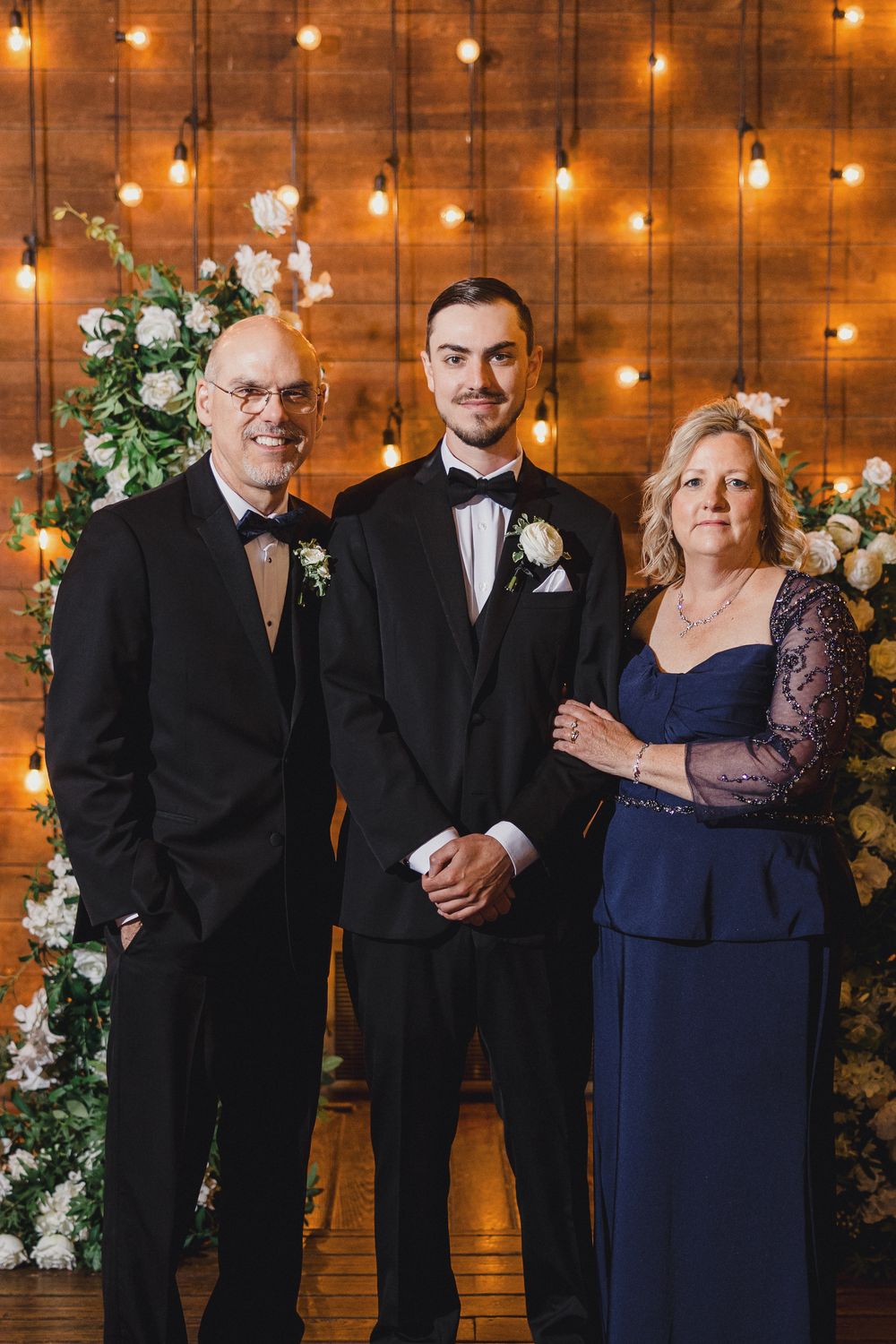 Wedding party in formal attire poses against rustic wood wall with glowing string lights and white floral decorations.
