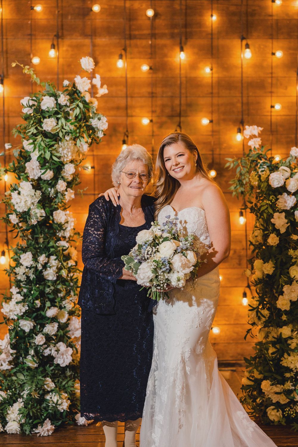 A wedding celebration features an elegant floral arch display with white roses and twinkling string lights against a wooden backdrop.