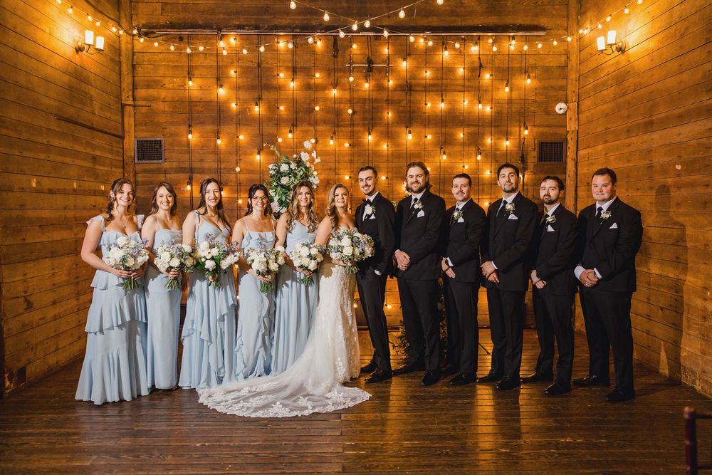 Wedding party poses in rustic barn venue with wooden walls and twinkling string lights backdrop.