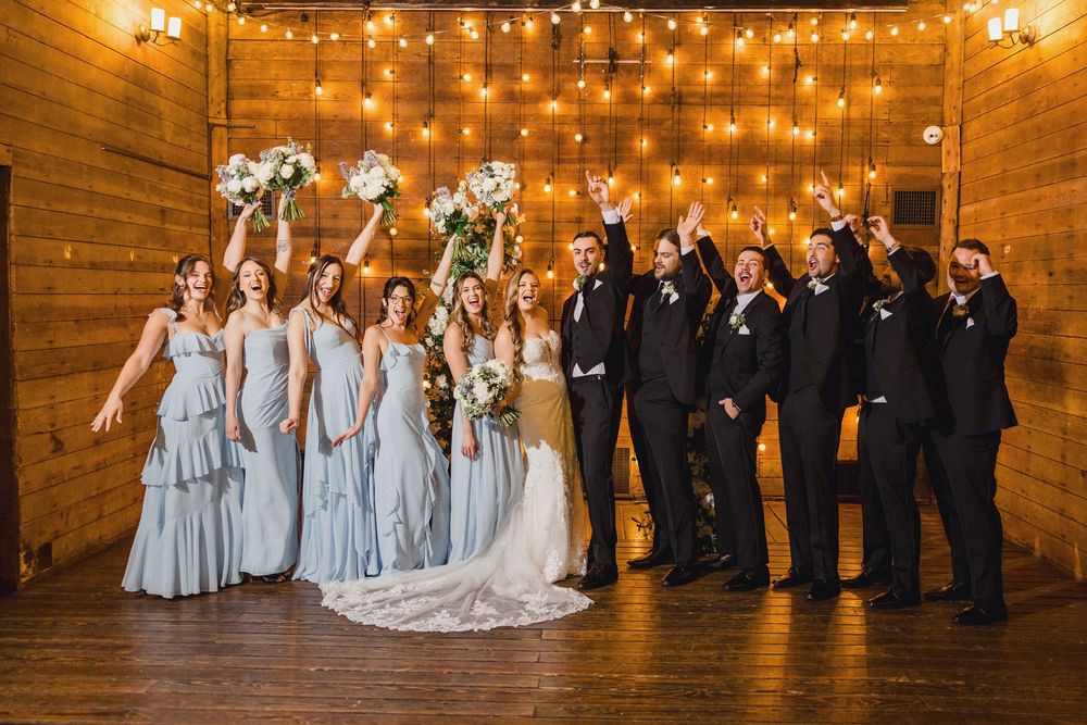 Wedding party in light blue dresses and black suits celebrating under string lights in a rustic wooden venue.