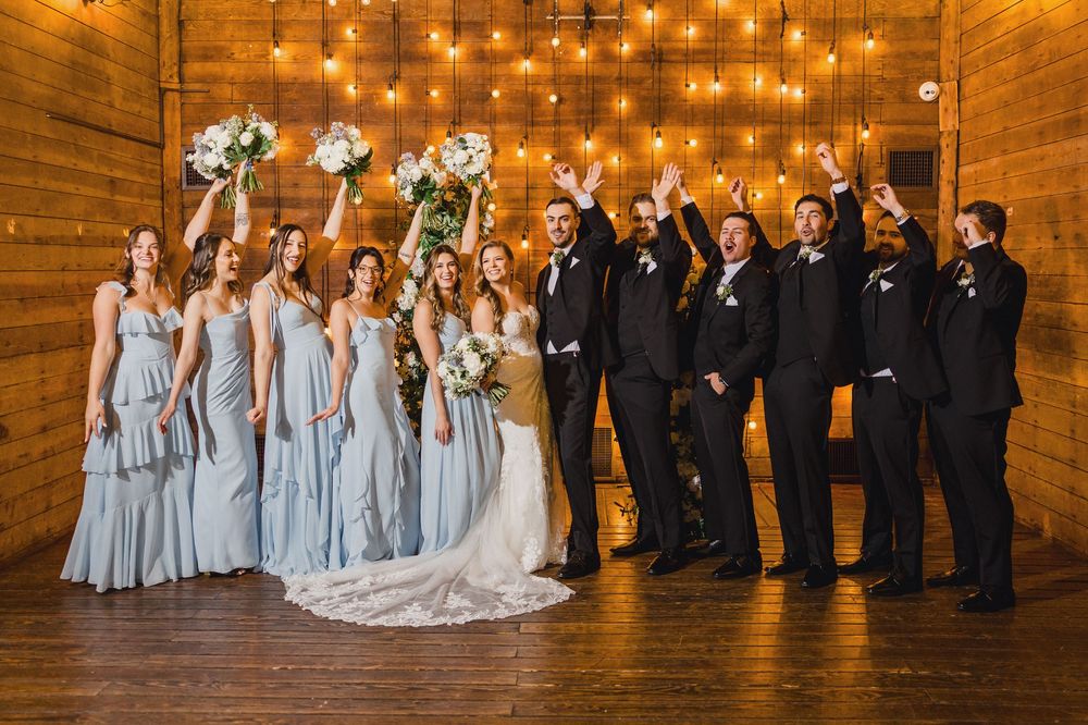 Wedding party poses together in rustic barn venue with string lights and wooden walls.