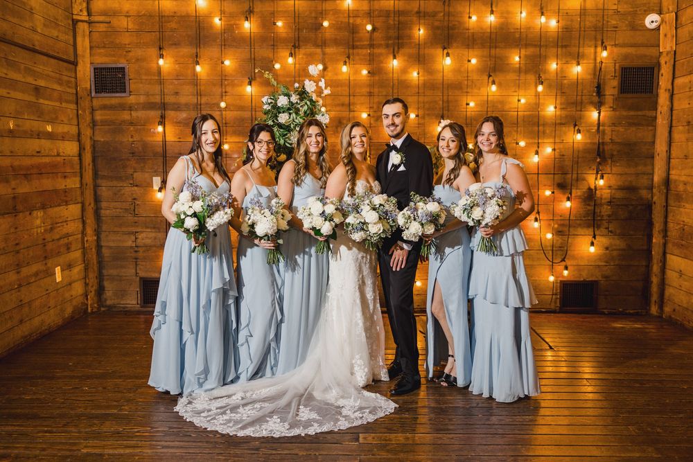 Bridal party in light blue dresses poses against warm wooden wall with string lights.