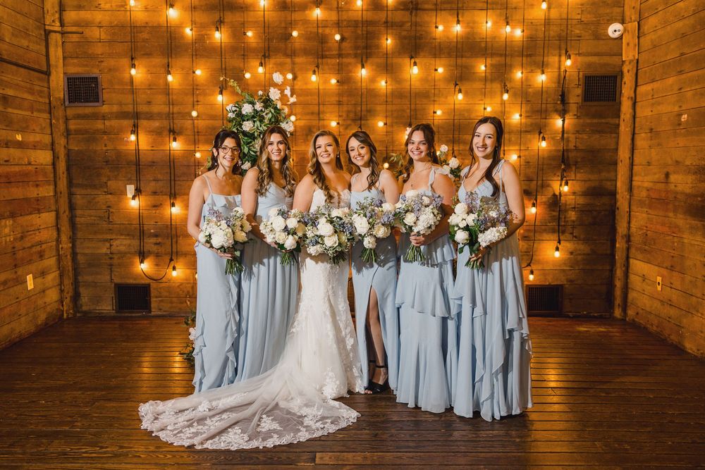 Wedding party poses in rustic barn venue with warm string lights and bridesmaids in light blue dresses.