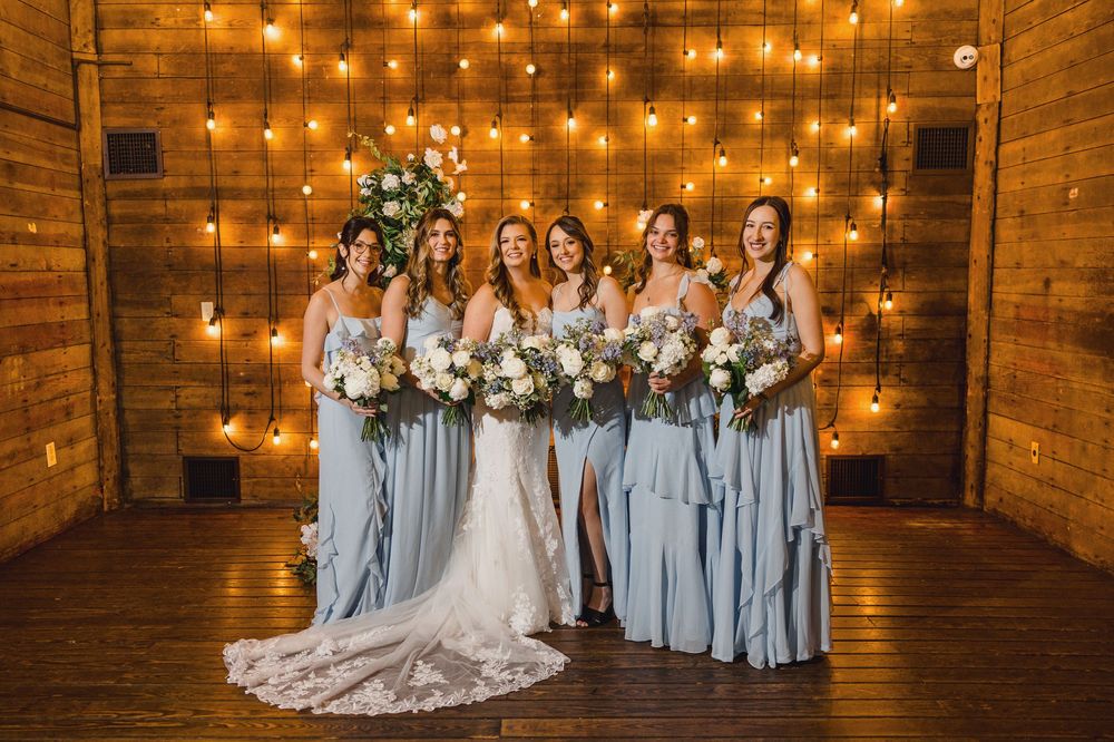 Bridesmaids in flowing blue gowns hold white bouquets against illuminated rustic backdrop.