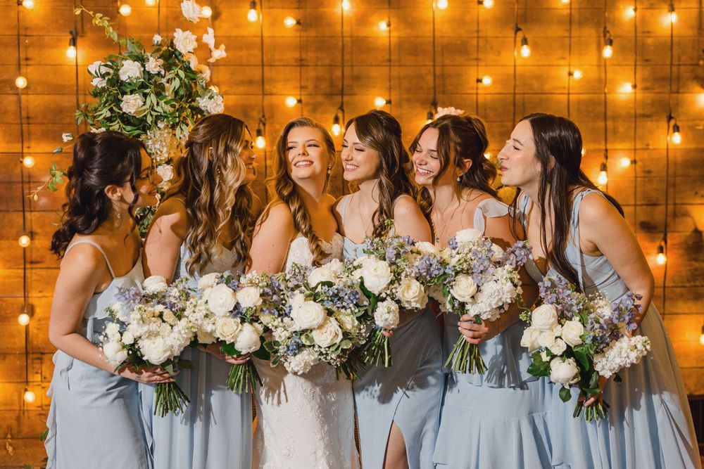 Wedding party in light blue dresses and black tuxedos pose with floral bouquets against rustic barn backdrop.