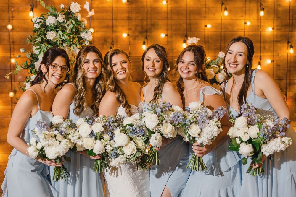Bridesmaids in light blue dresses hold white bouquets against a warmly lit wooden backdrop.