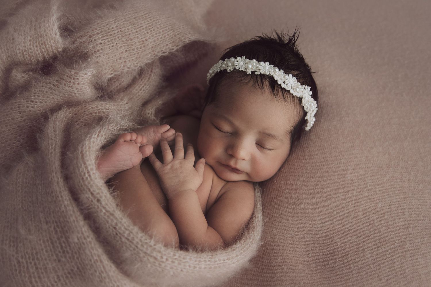 Sleeping newborn baby wearing white lace headband swaddled in soft pink blanket in professional studio setting.