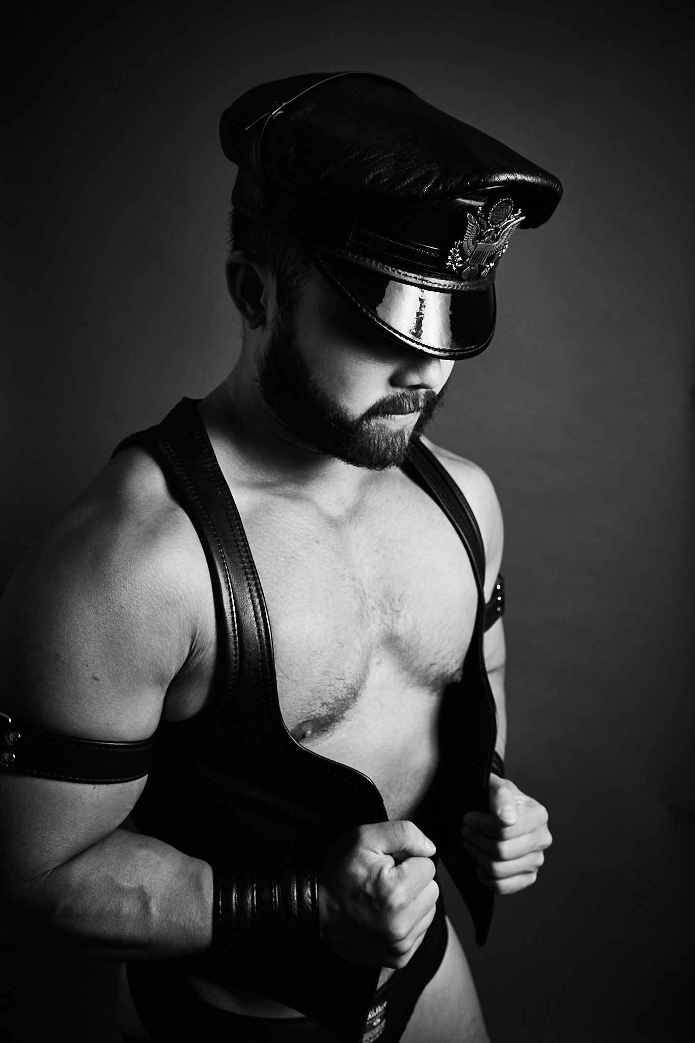 Muscular bearded man in leather cap and harness poses in dramatic black and white studio portrait.