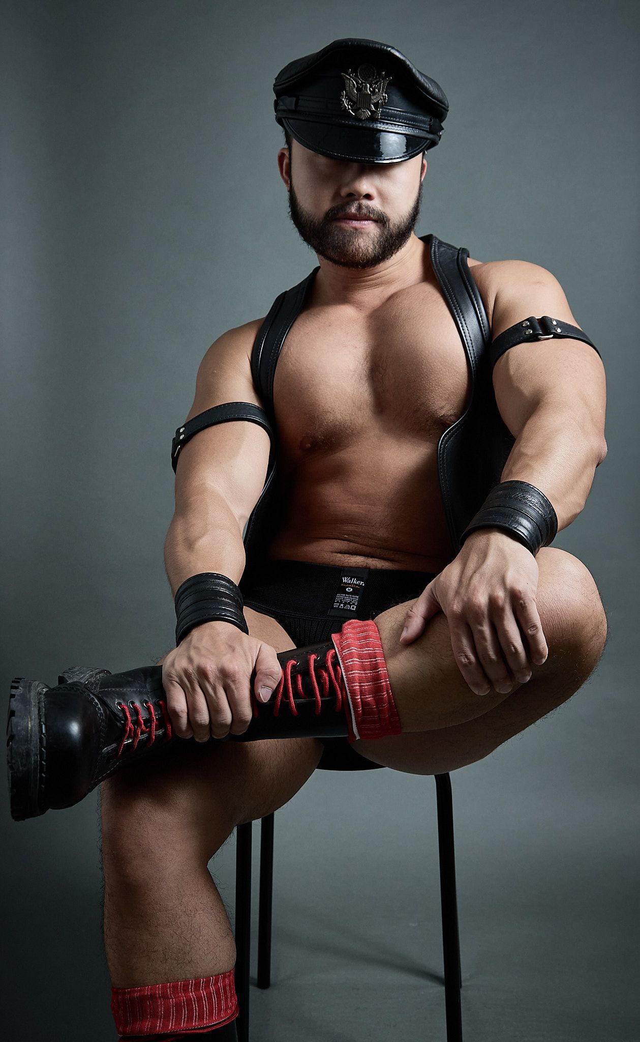 Muscular bearded man in leather cap and harness sits on stool holding red laced boot in studio photo.