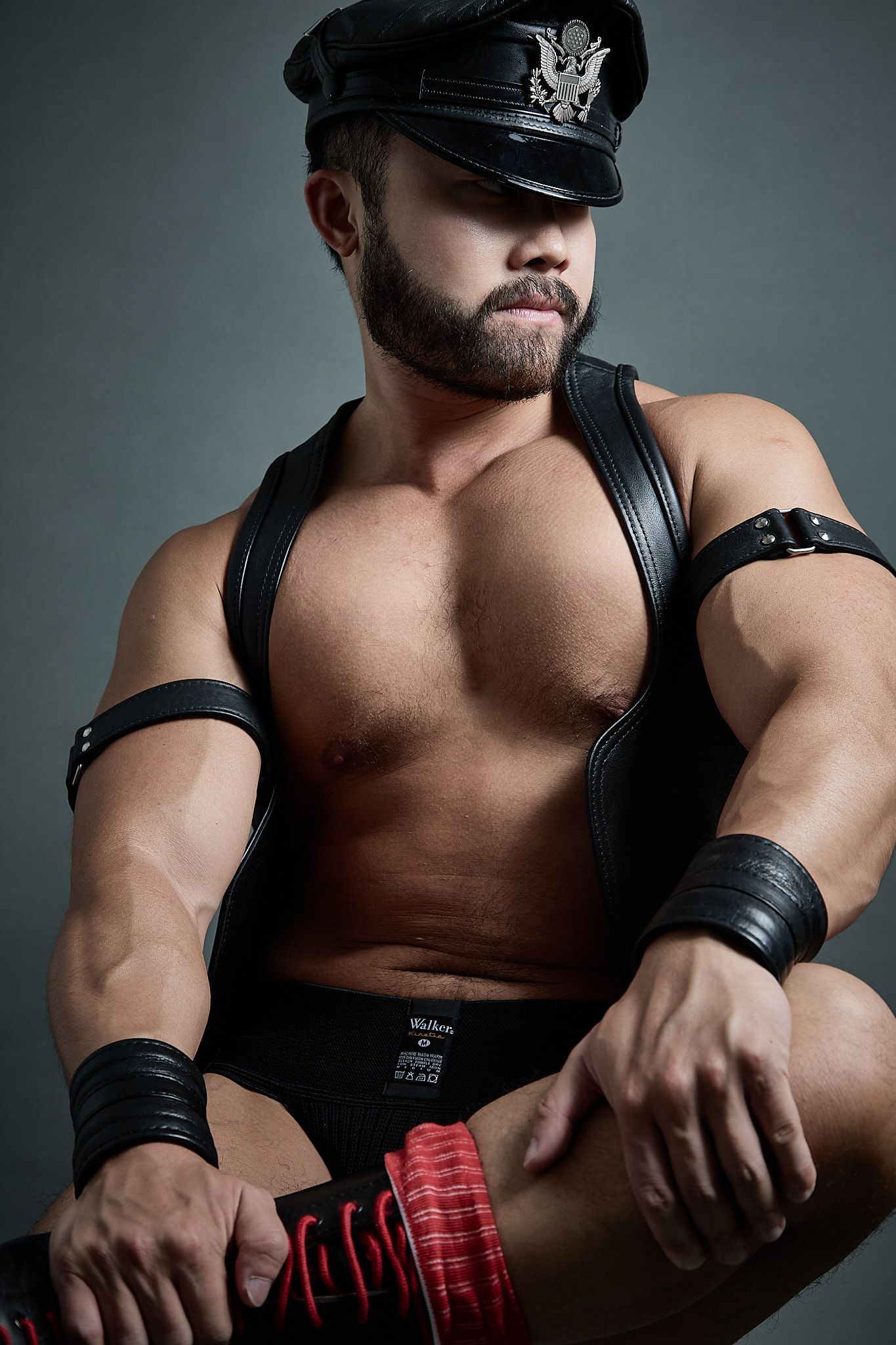 Muscular bearded man in black leather cap, harness, and wrist cuffs poses in dramatic studio lighting.