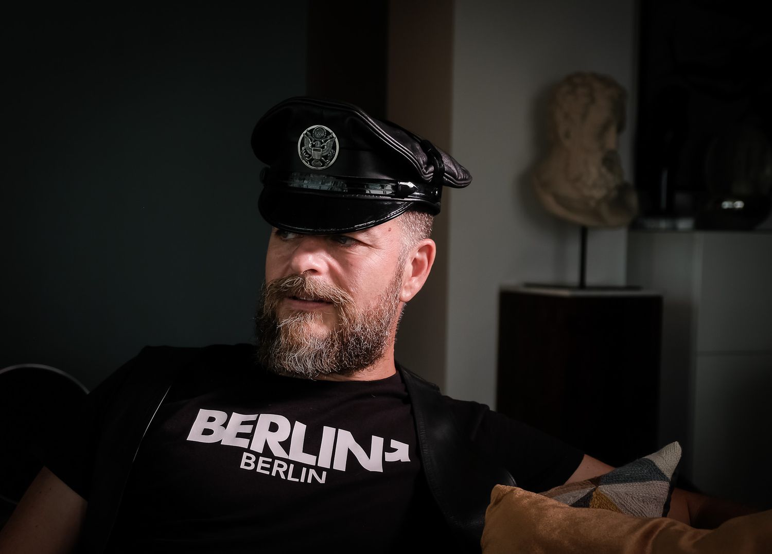 Bearded man wearing a black leather cap and Berlin t-shirt sitting indoors.