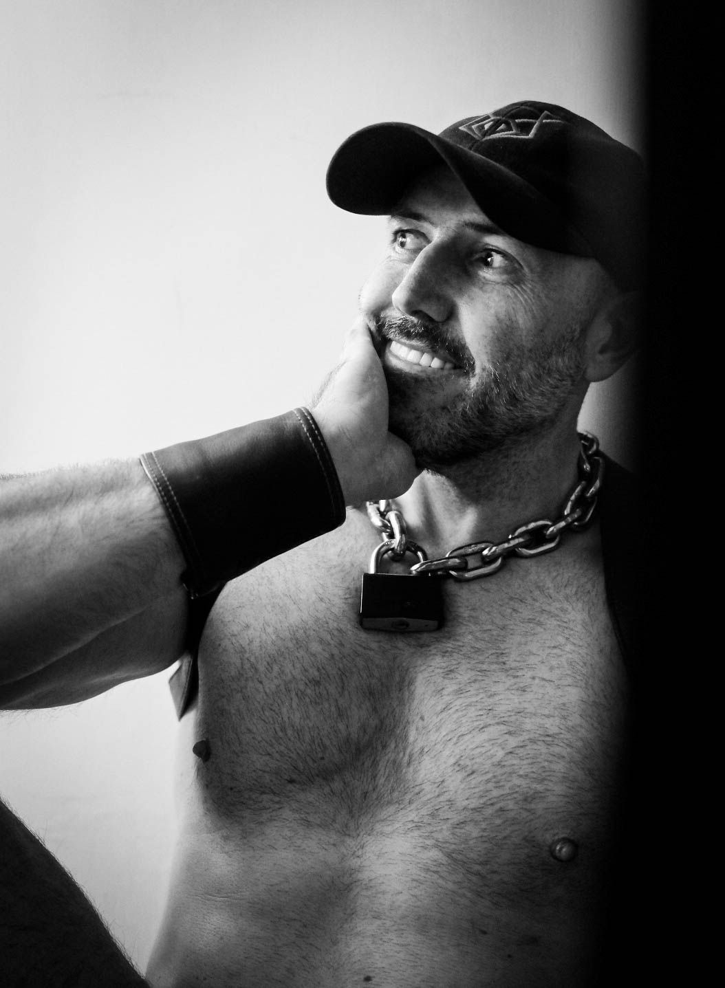 Muscular shirtless man wearing cap and chain necklace smiles upward in black and white portrait.