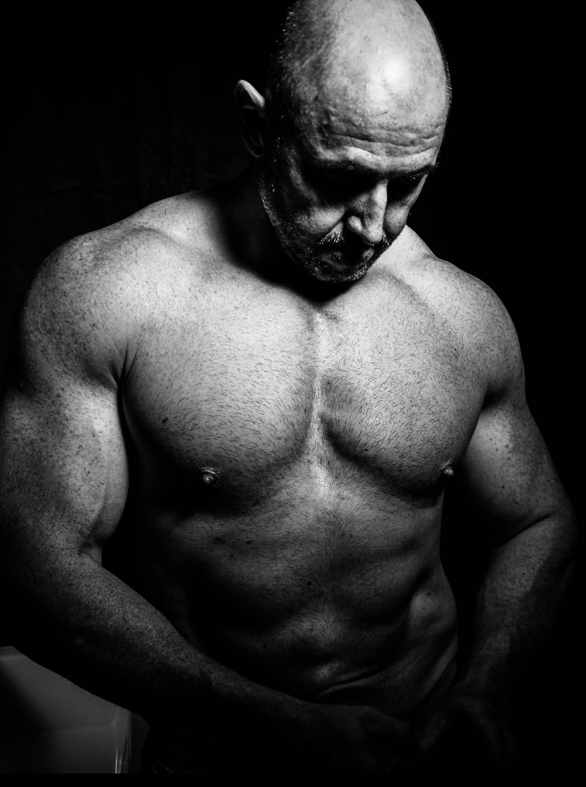 Bald muscular shirtless man flexes in a dramatic black and white portrait with dark background.