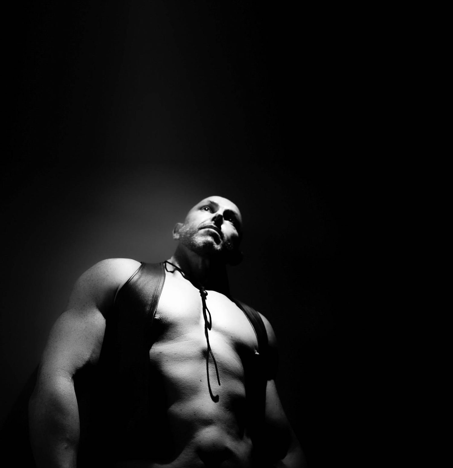 Muscular shirtless man with harness gazes upward in dramatic black and white low-key lighting.