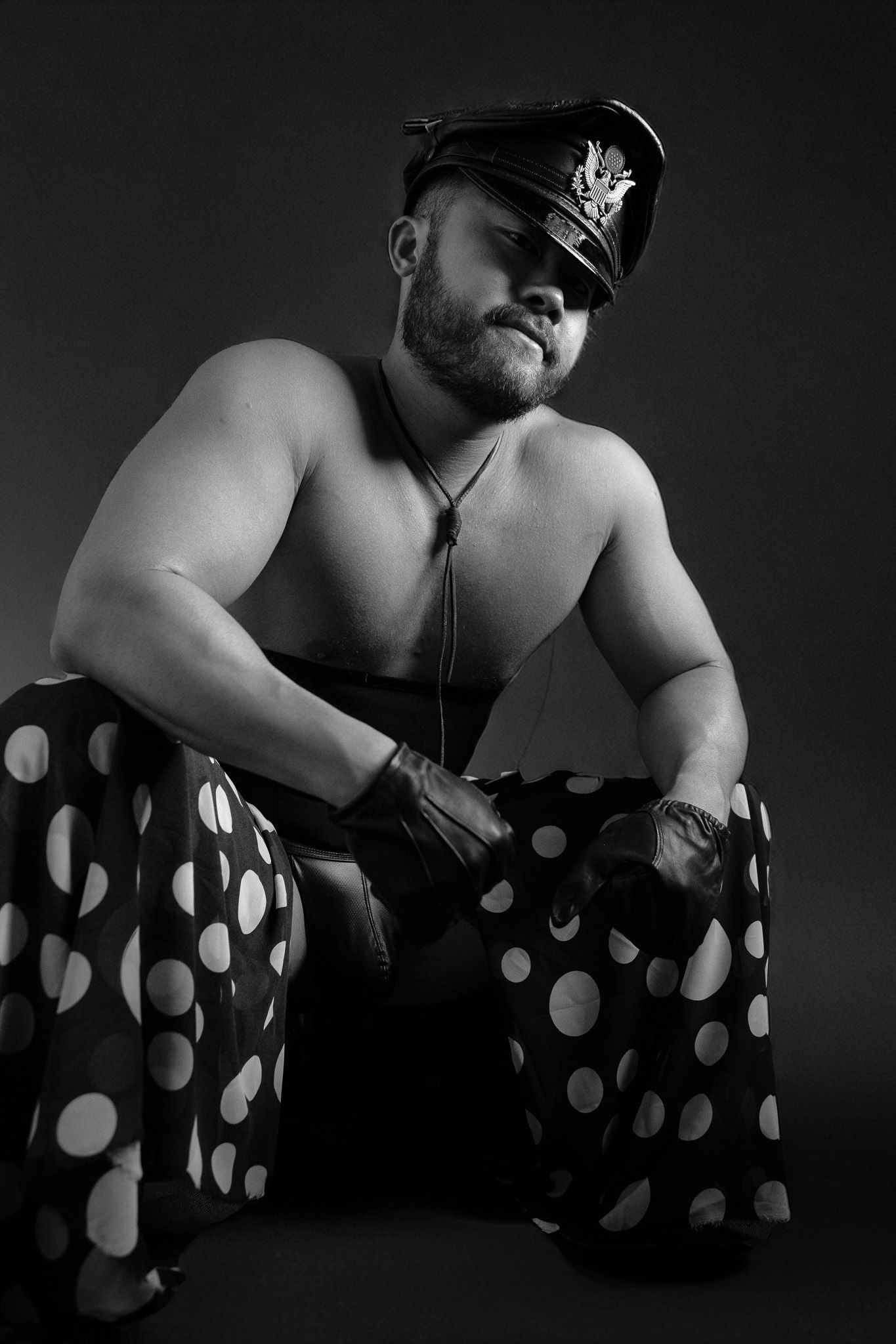 Muscular shirtless man in black leather cap and polka dot pants poses in dramatic black and white studio photo.