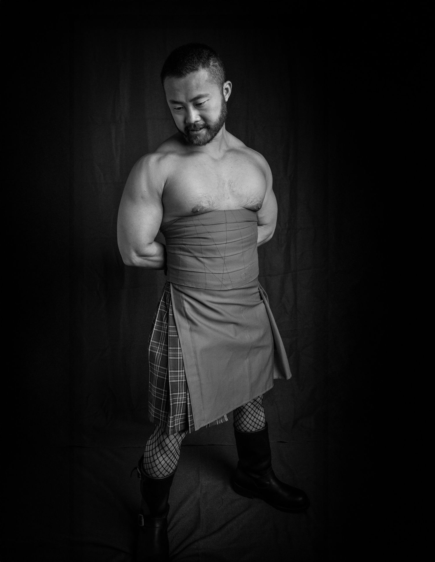 Muscular shirtless Asian man wearing plaid kilt, fishnet stockings, and boots in black and white studio portrait.