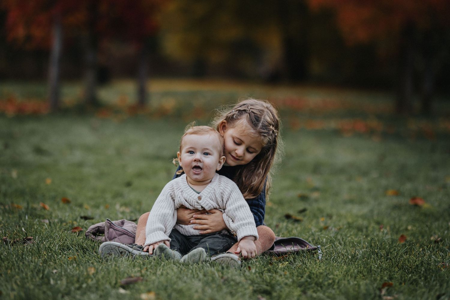 Two figures share a joyful moment together while sitting on grass during autumn.