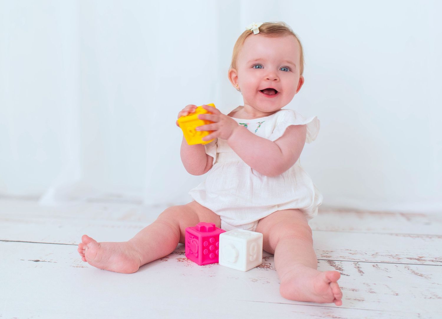 Baby in white outfit playing with colorful blocks on wooden floor.