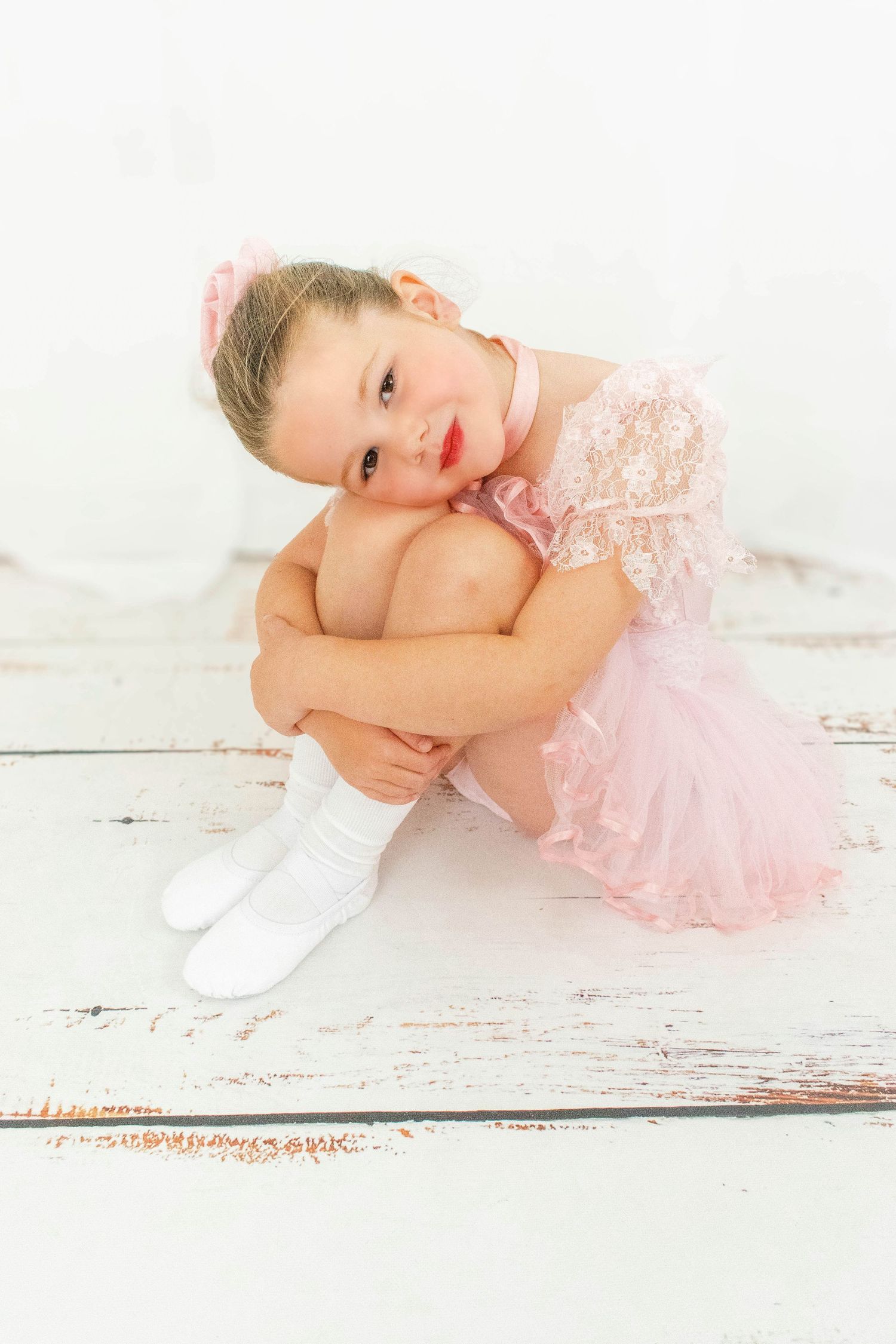 Young dancer in pink tutu poses gracefully on white wooden floor.