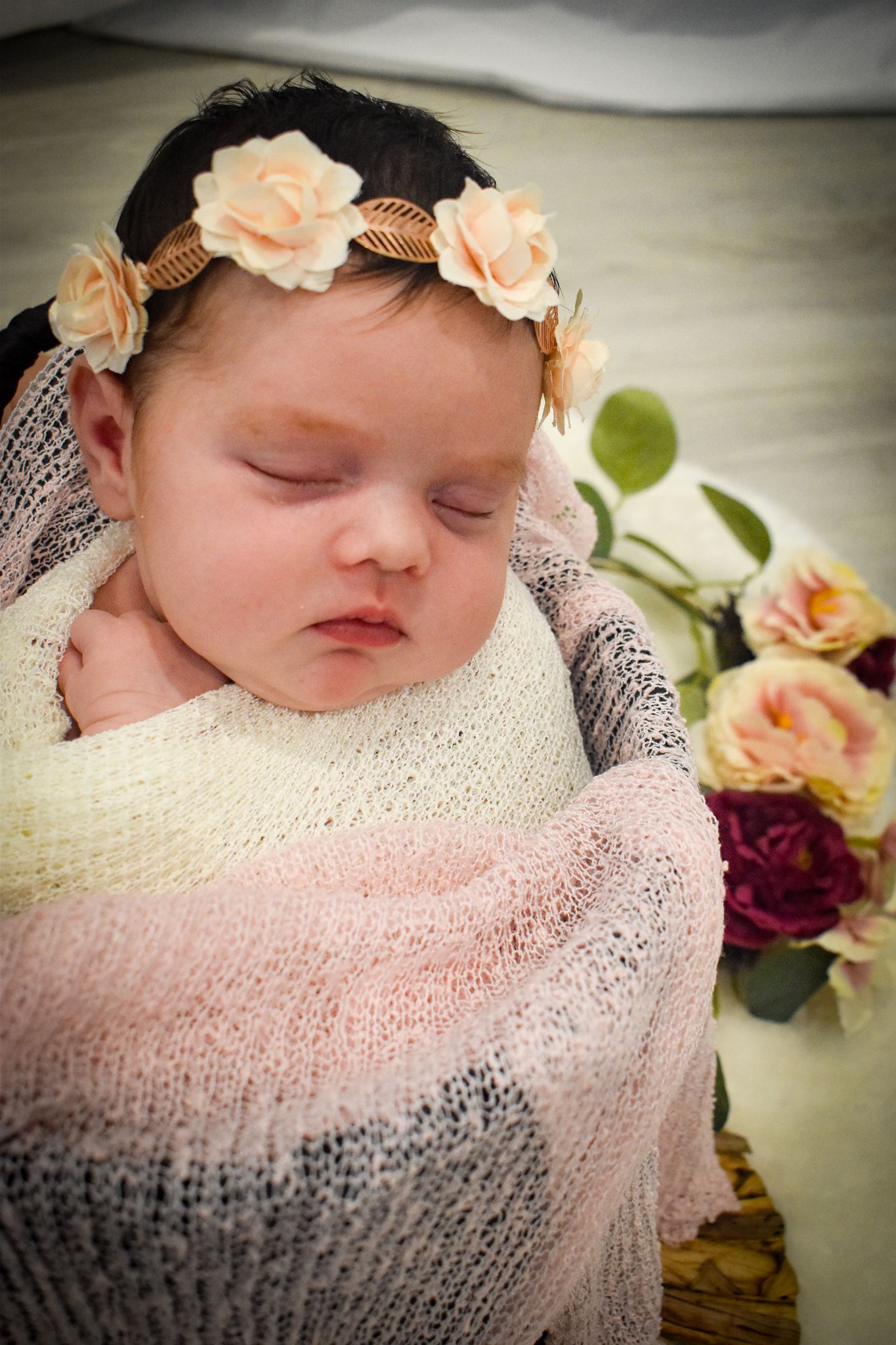 Sleeping newborn wearing floral headband wrapped in pink and gray knit blanket.
