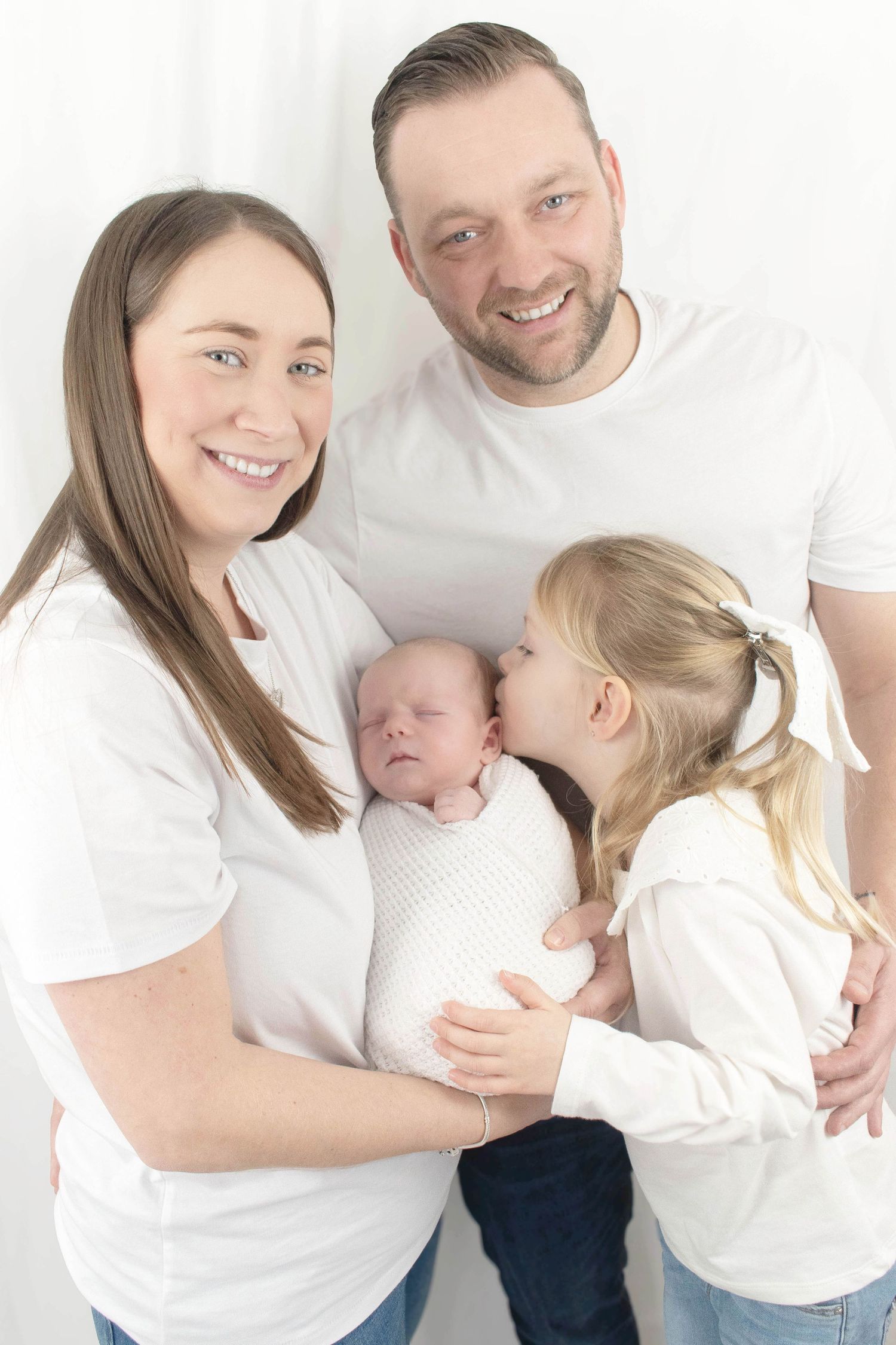 Family portrait with newborn baby in white outfits against light background.