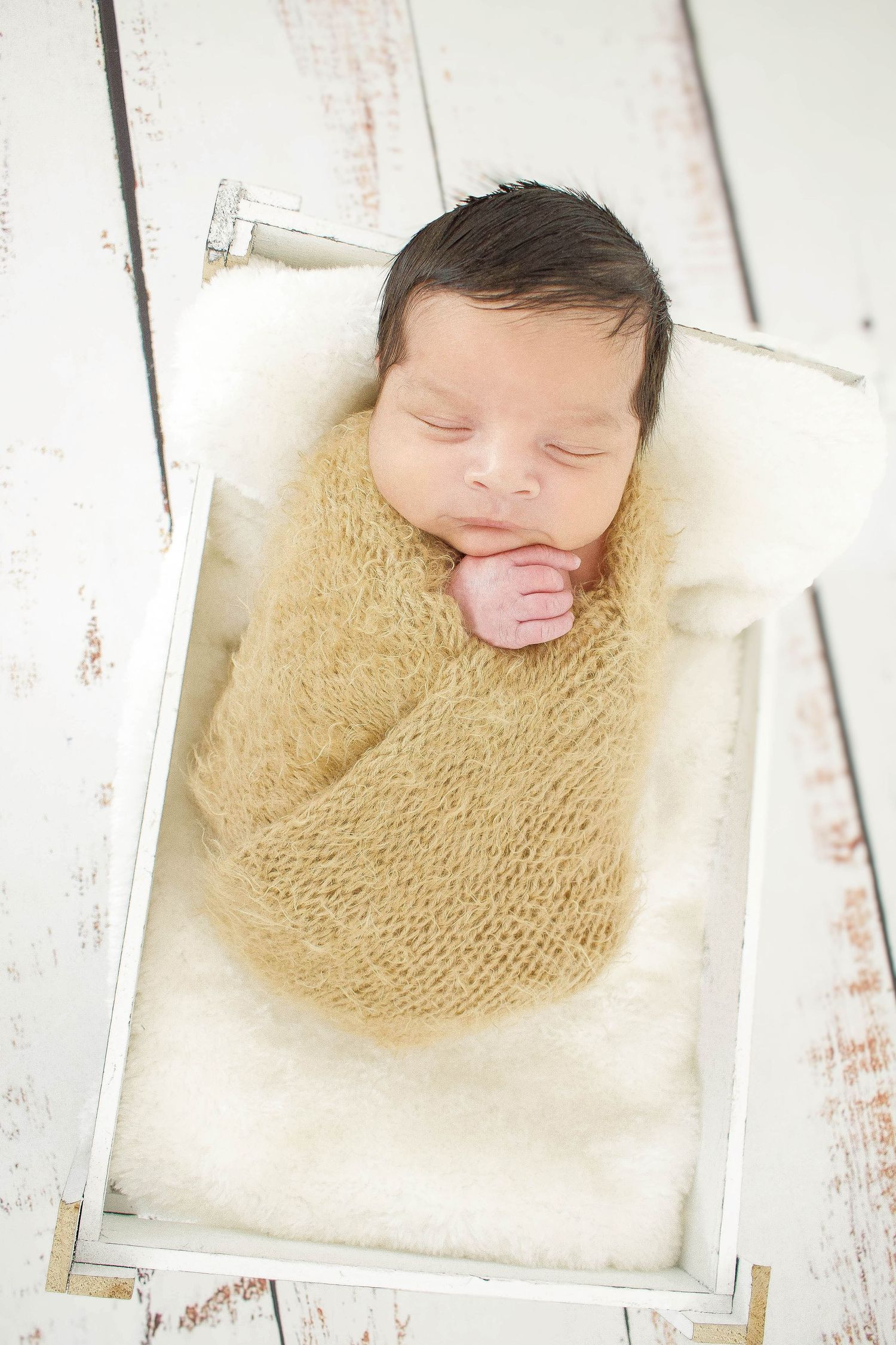 Sleeping newborn wrapped in yellow knit blanket lying in white wooden box.