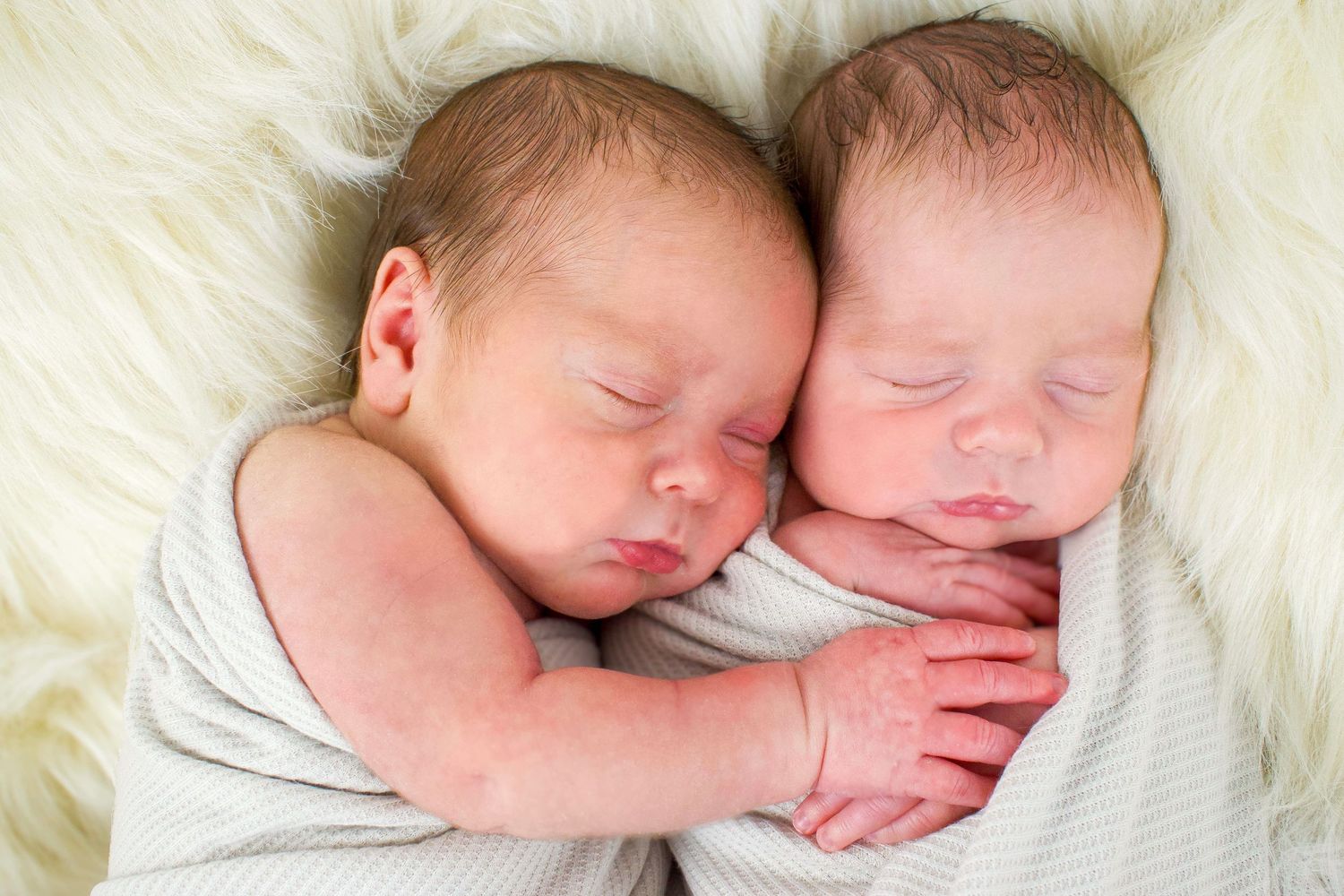 Two sleeping newborn babies swaddled together on white fur background.