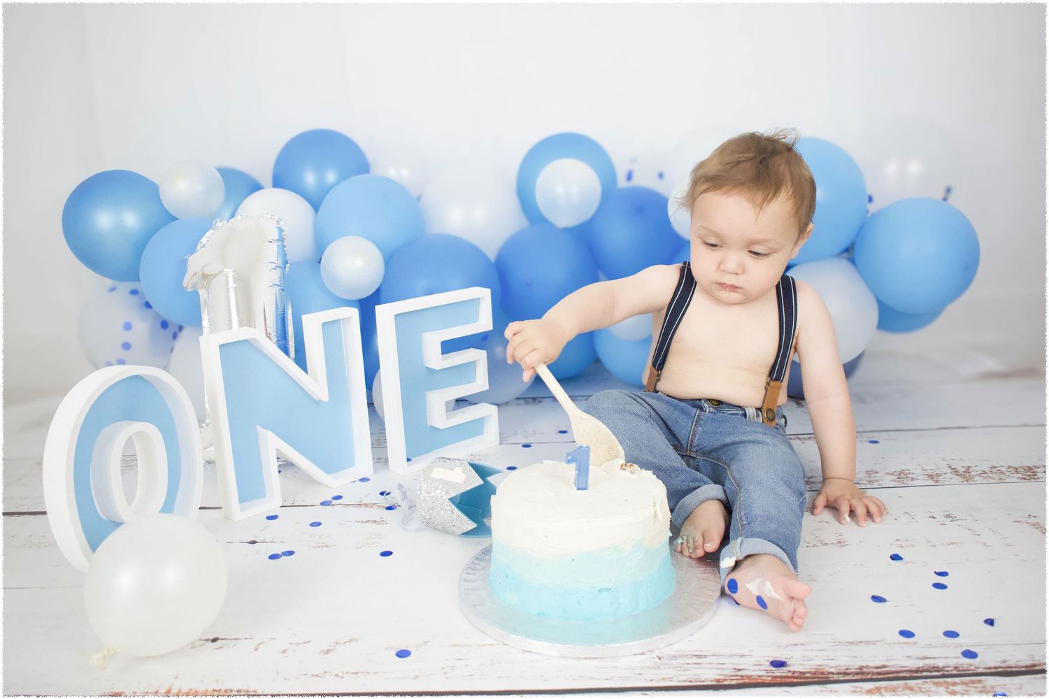 First birthday celebration setup with blue balloons, 'ONE' lettering, and a smash cake on white background.