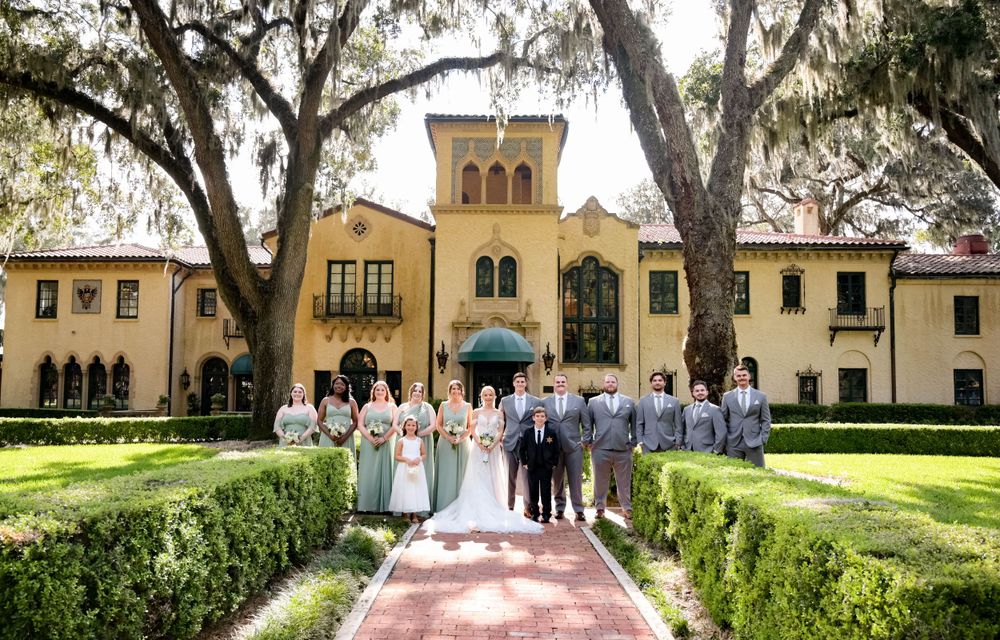 Wedding party poses in front of elegant Spanish-style yellow mansion with manicured gardens and oak trees.