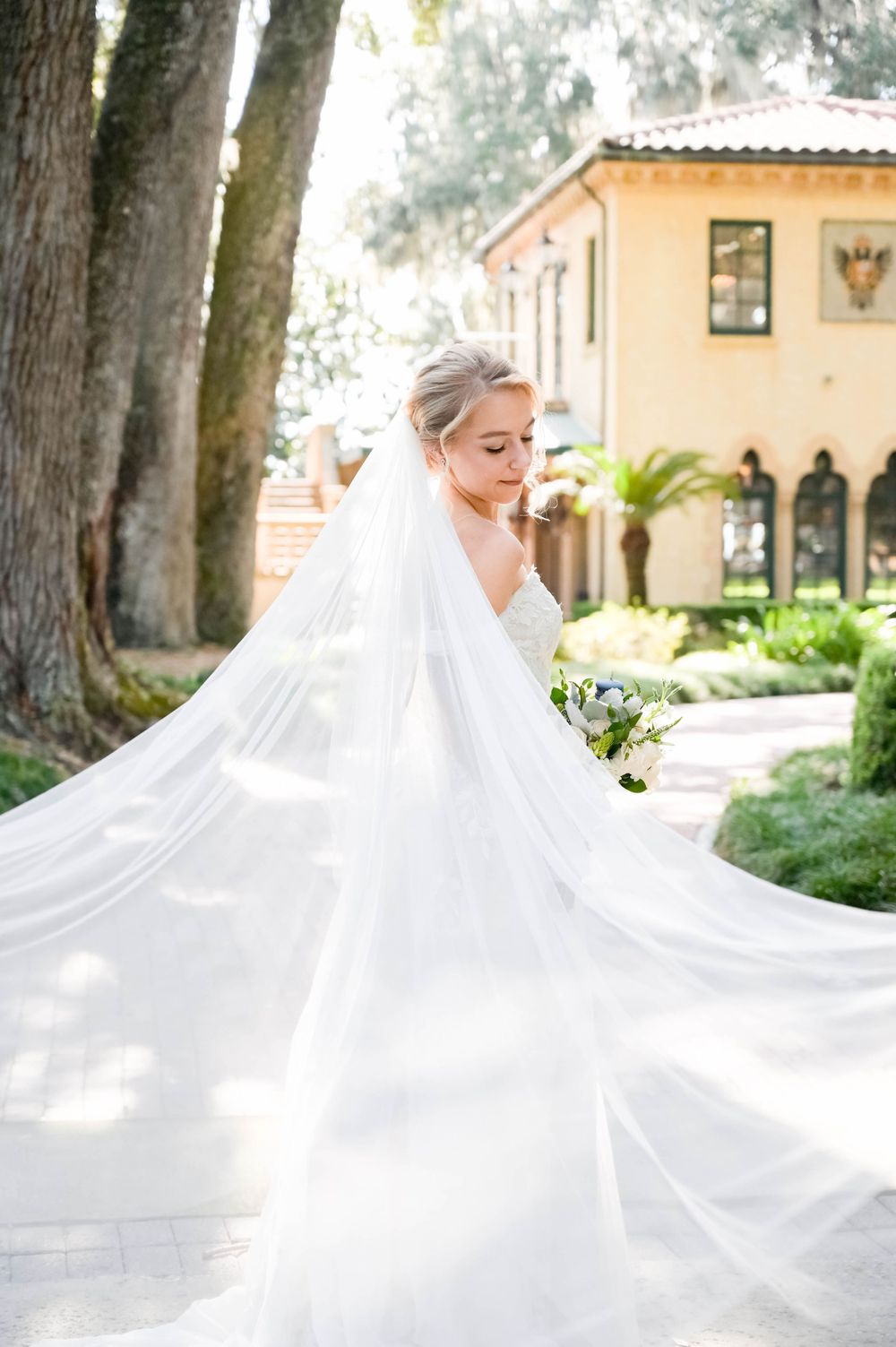 A flowing white wedding dress and veil catch the light in a dreamy garden setting.