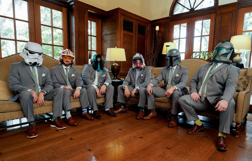 Groomsmen wearing Star Wars stormtrooper masks pose in matching gray suits in a wood-paneled room.