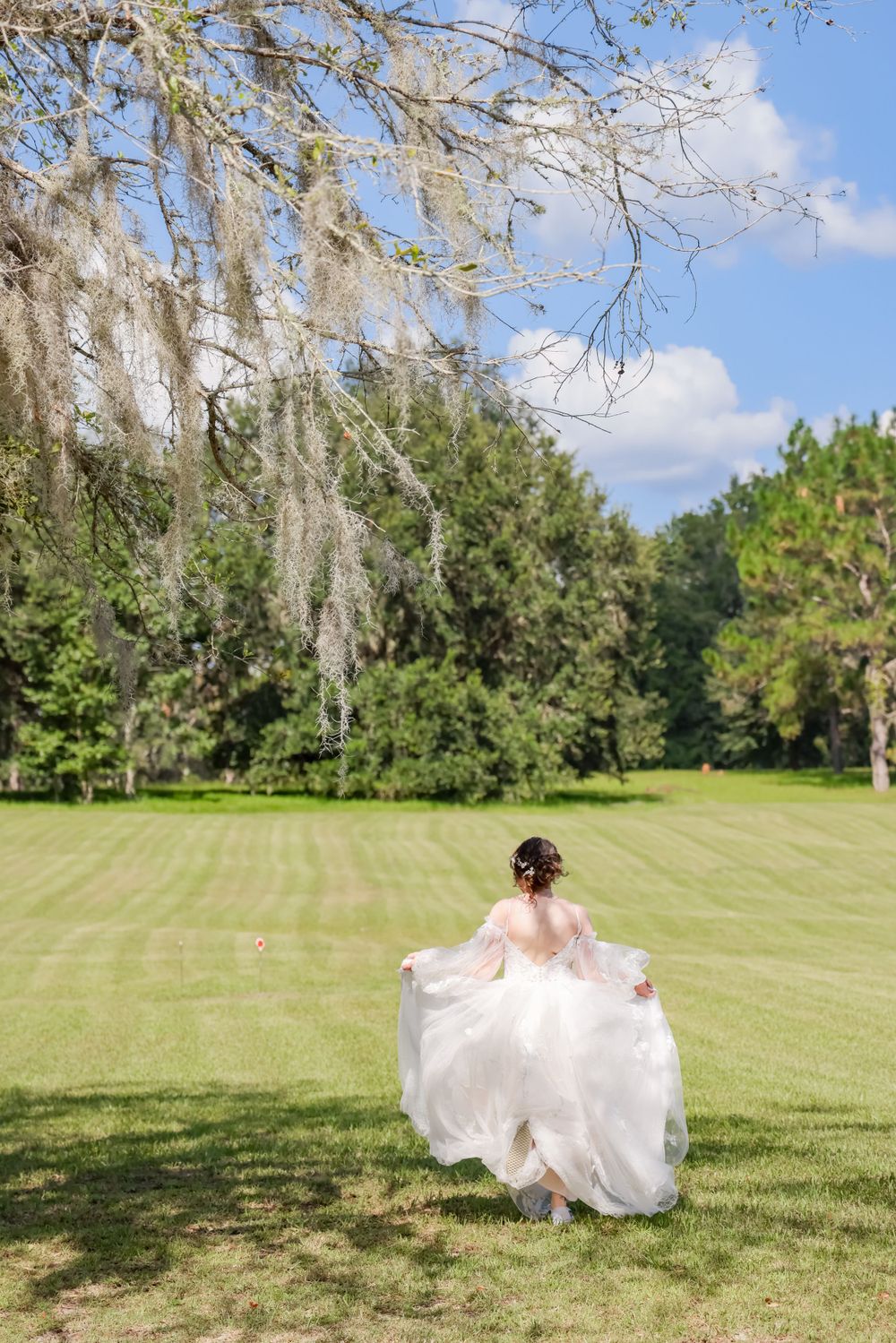 Bride in flowing white dress twirling on manicured lawn beneath Spanish moss-draped trees.