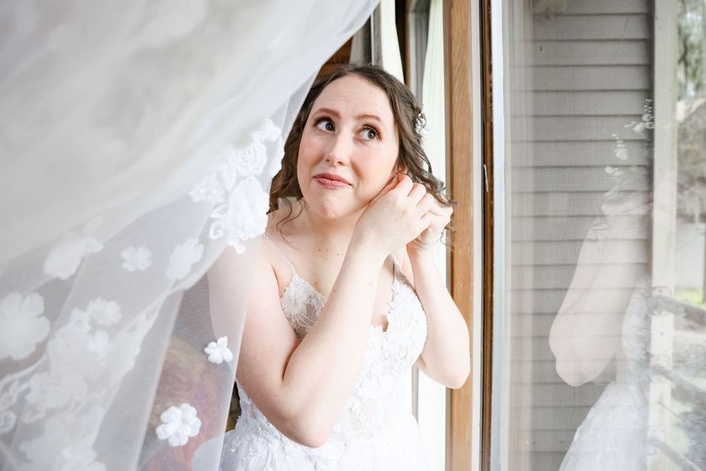 A person in a white lace wedding dress adjusting earrings near a window.