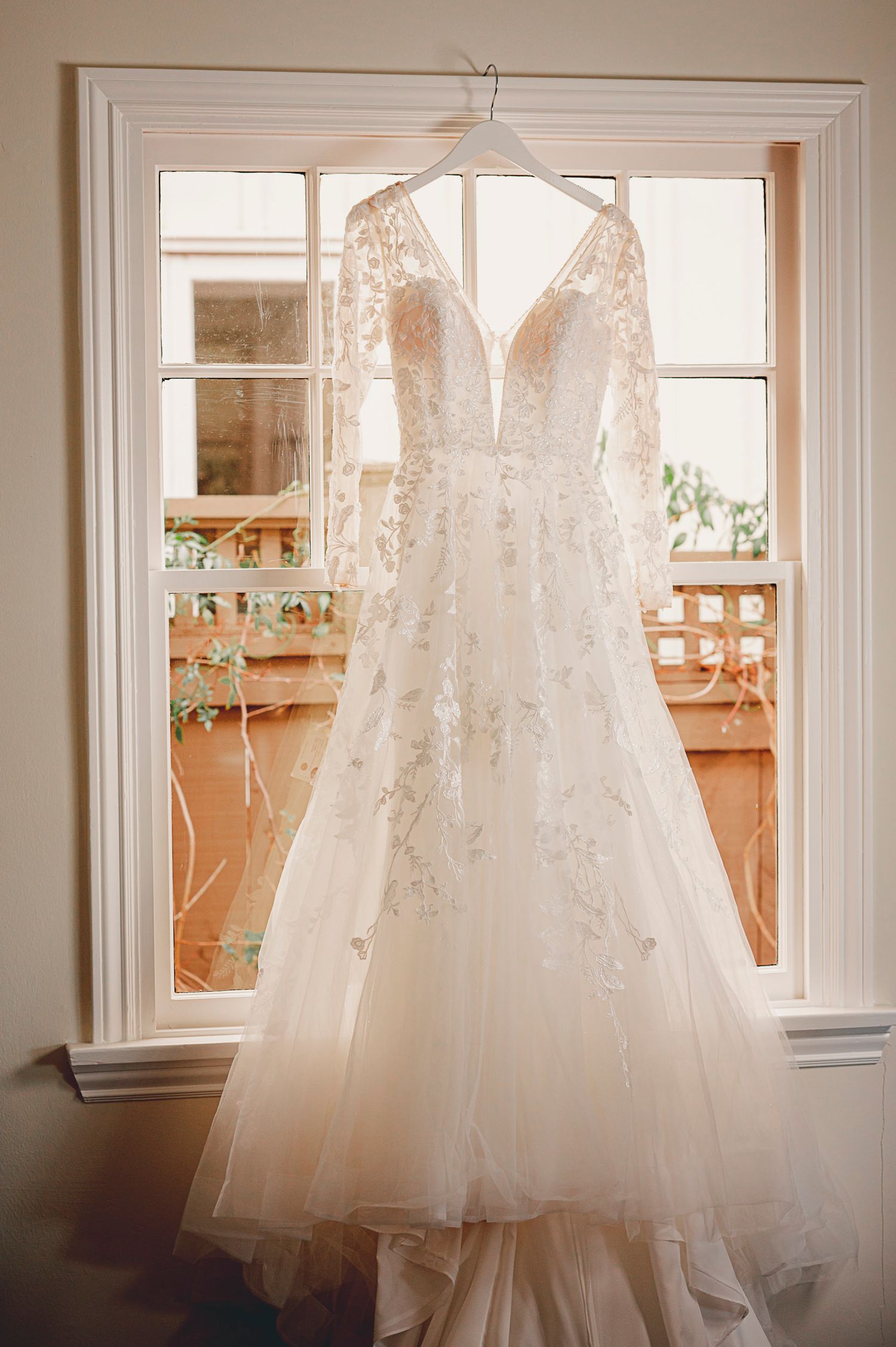 Elegant white lace wedding dress hanging in front of a sunlit window.