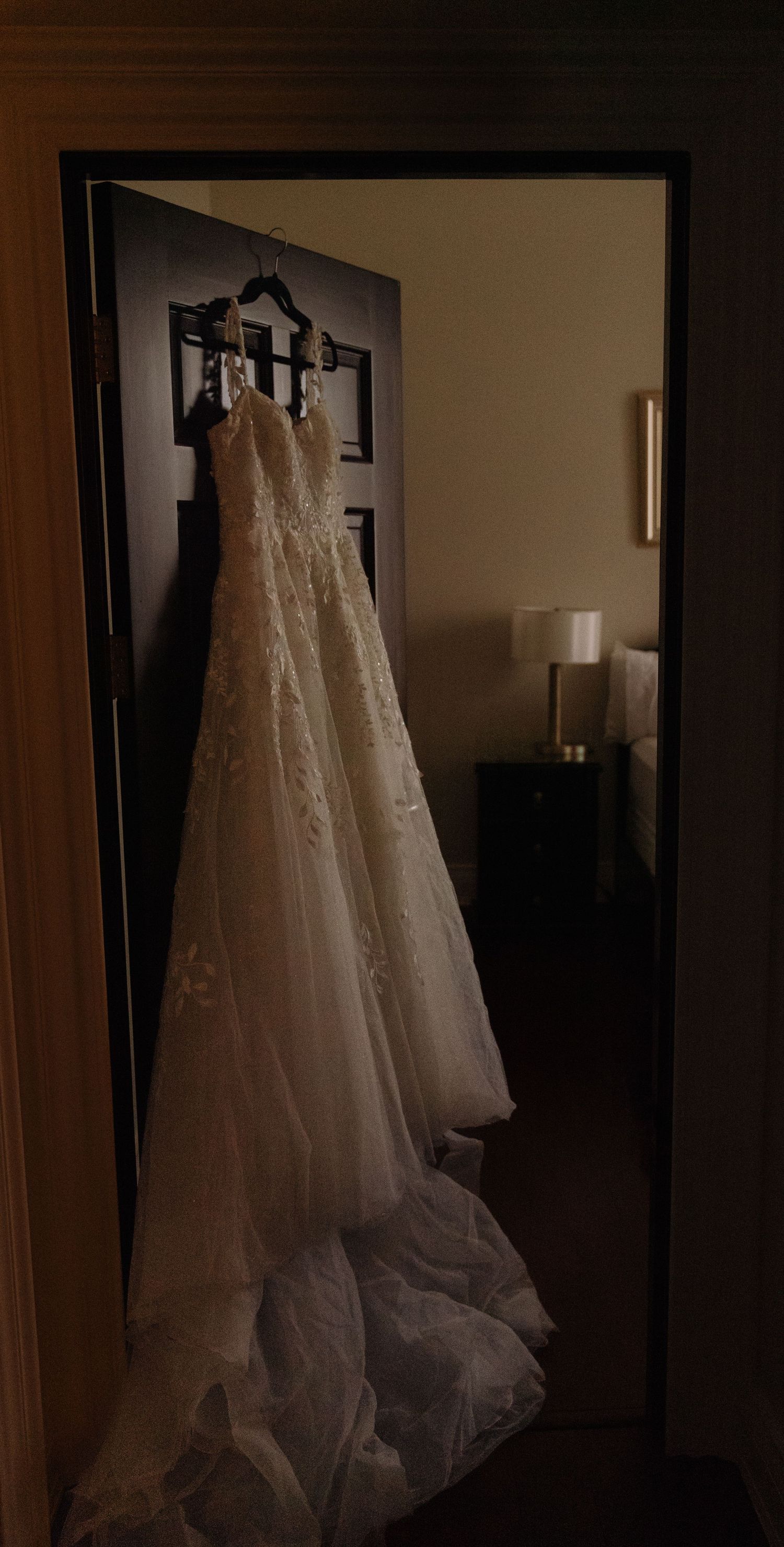 A white lace wedding dress with a long train hangs elegantly on a dark door in a dimly lit room.