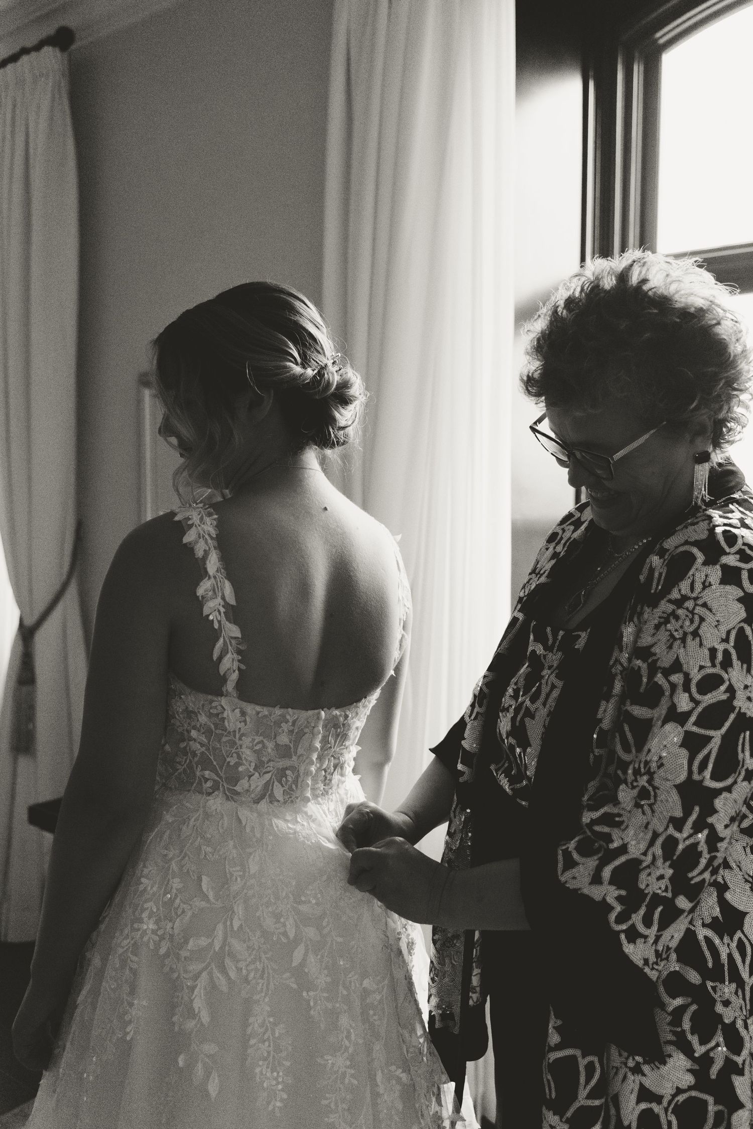 A black and white photo captures the intricate back details of a wedding dress being adjusted.