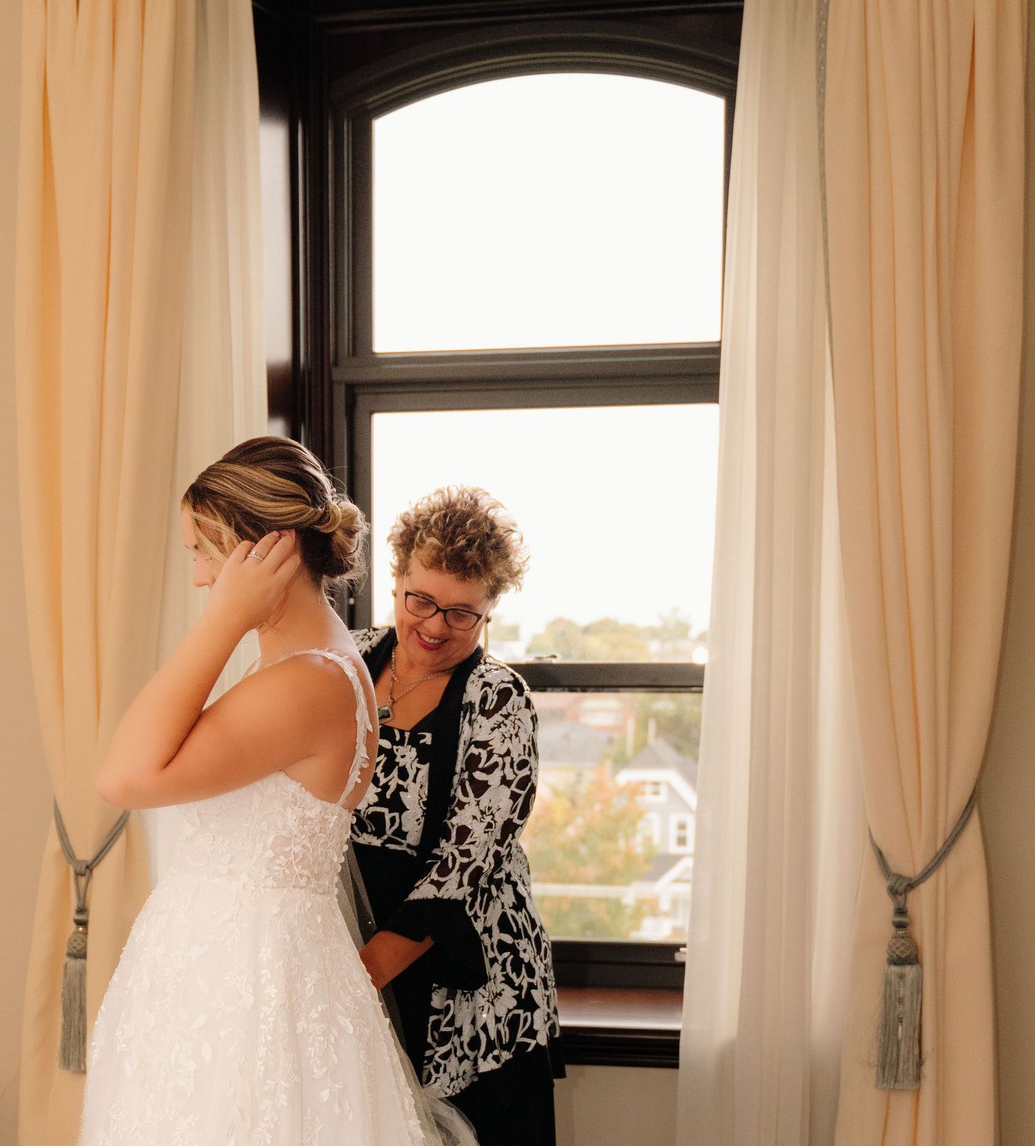 A sunlit window frames a tender moment during wedding dress preparations.