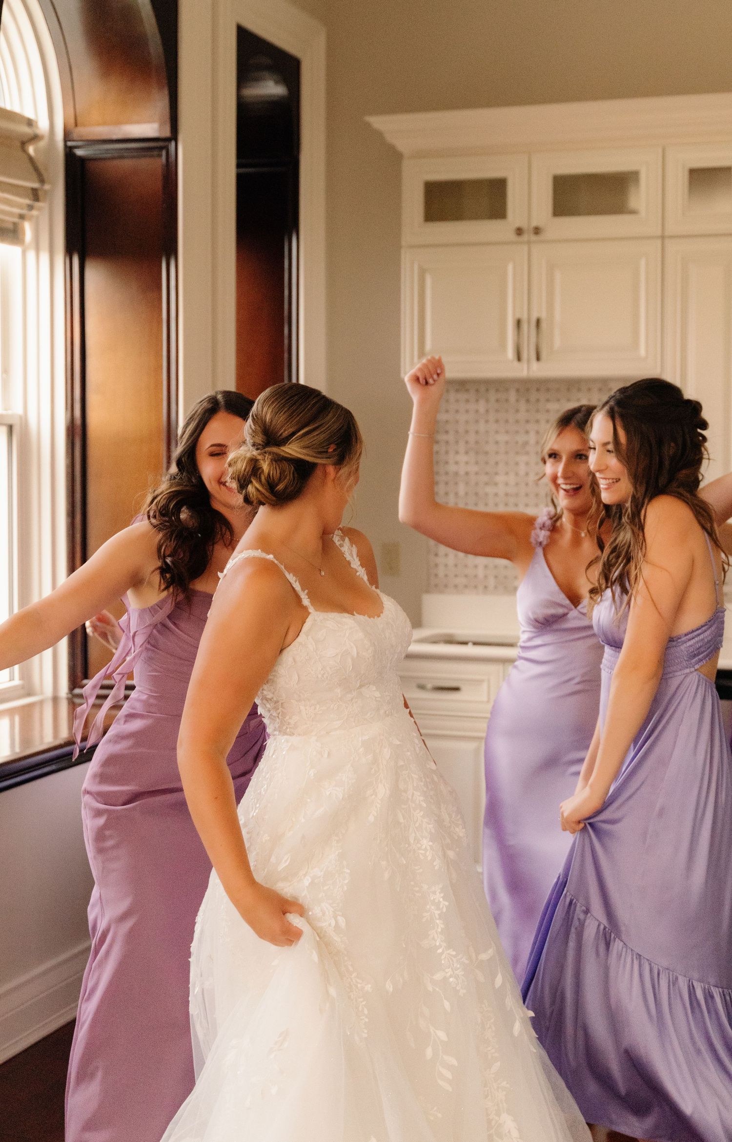 Wedding party celebrating in a bright kitchen while wearing purple bridesmaid dresses.