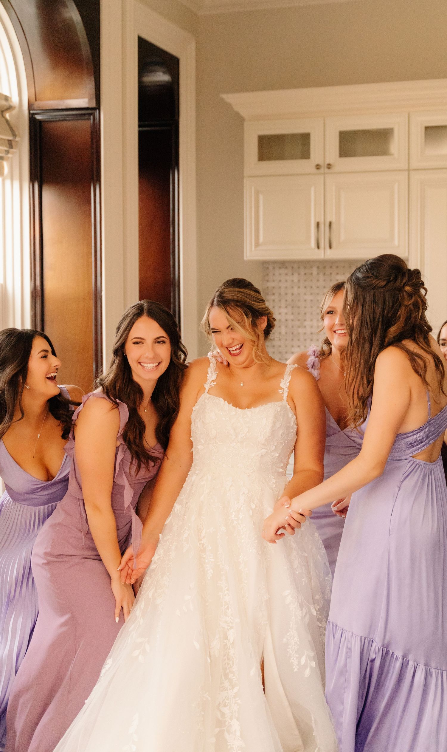 Bridal party in lavender dresses sharing a joyful moment in an elegant kitchen setting.